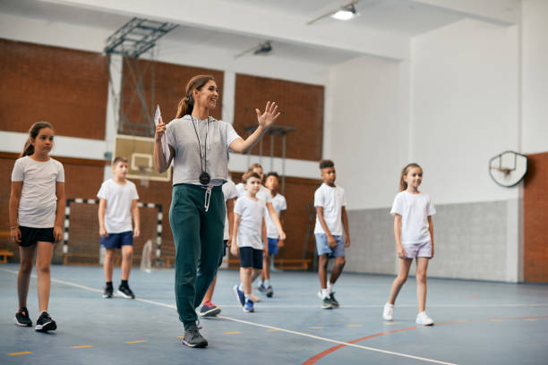 Young female coach having PE class with group of elementary students at school gym. Happy physical education teacher talking to her students during a class at elementary school gym. physical education stock pictures, royalty-free photos & images