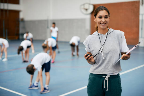 Portrait of happy female physical education teacher at school gym. Young happy coach using stopwatch during PE class at school gym and looking at camera. Her students are exercising in the background. school gymnasium stock pictures, royalty-free photos & images