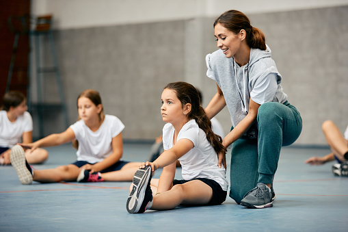 Little girl stretching on the floor and warming up with help of PE teacher during a class at school gym.