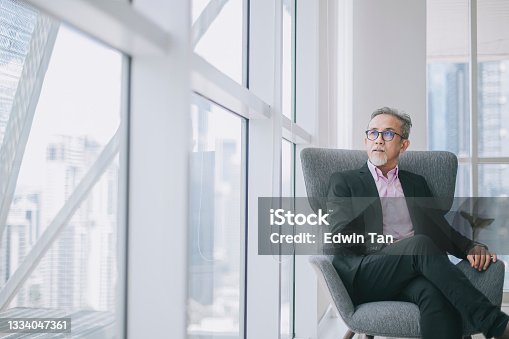 istock Successful Asian chinese senior businessman in suit sitting crossed leg on sofa in his office room looking at the capital city 1334047361