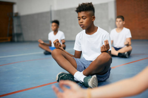 Black elementary student doing breathing exercise while practicing Yoga on physical education class. African American schoolboy and his friends meditating in lotus position during PE class at school gym. mindfulness children stock pictures, royalty-free photos & images