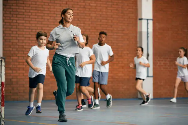 Photo of Physical education teacher and group of elementary students running while warming up at school gym.