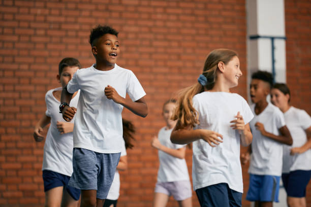 Black elementary student and his friends running on physical education class at school gym. Multi-ethnic group of school children running on PE class at school gym. Focus is on African American boy. physical education stock pictures, royalty-free photos & images