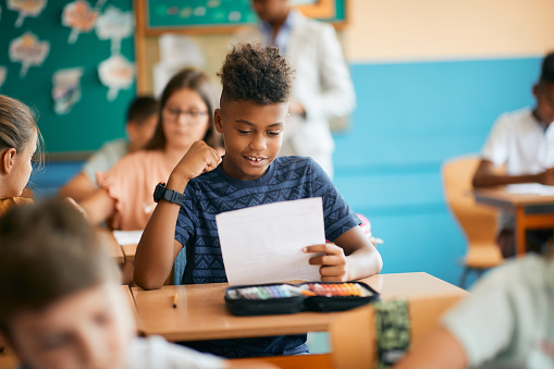 Happy black elementary student celebrating getting a good grade in the classroom at school.