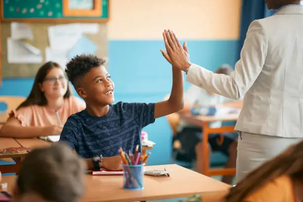 Photo of Happy black elementary student and his teacher giving high five during class at school.