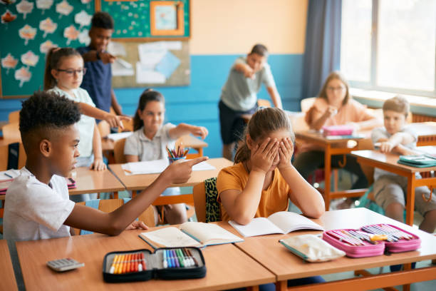 Group of elementary school kids mocking their classmate in the classroom. Small girl feeling upset while her classmates make fun of her during a class at elementary school. teasing stock pictures, royalty-free photos & images