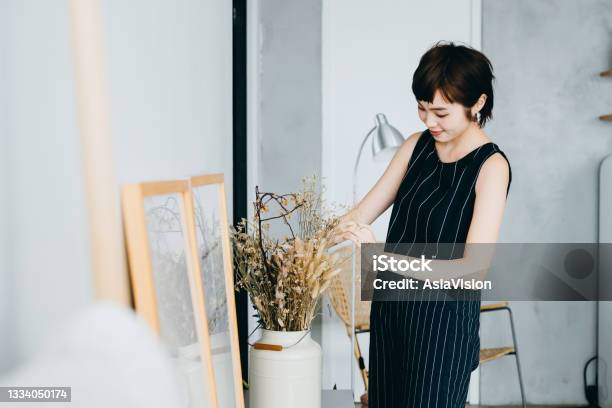 Young Asian Woman Decorating And Arranging A Vase With Dried Plants On Cabinet In The Living Room At Home Stock Photo - Download Image Now