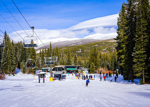 View of chairlift at the bottom of ski slope in Winter Park, Colorado, ski resort; skiers and snowboarders waiting their turn to go up  on a cold winter day; mountain range in the background