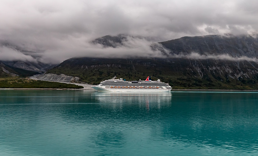 Alaska, USA - August 23 , 2018: Cruise ship Carnival Splendor sailing in one of Alaskan fjords. Massive mountains covered with clouds in the background. Turquoise water in the foreground.