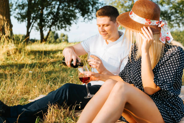 pique-nique romantique en plein air. beau jeune couple hétérosexuel en vacances dans la nature, l’homme verse du vin rouge dans un verre à sa petite amie au chapeau de paille et robe d’été à pois. histoire d’amour - women love heterosexual couple color image photos et images de collection