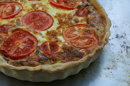 Photo showing elevated view of baking sheet containing a fluted pastry crust, homemade, quiche lorraine topped with cherry plum tomato halves.