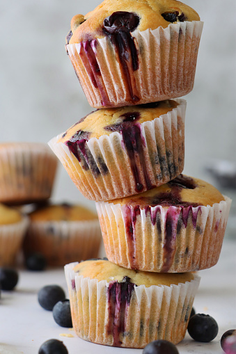 Stock photo showing close-up view of fresh blueberries surrounding a stack of homemade blueberry muffins in paper cake cases against a marble effect background.