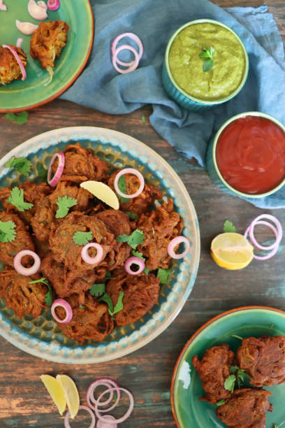 Stock photo showing an elevated view of a batch of homemade crispy onion bahjees in a pile. imagen de la pila de bhajis de cebolla crujiente caseros / pakoras aderezados con hojas de cilantro y anillos de cebolla roja en un plato estampado al lado de la muselina azul y cuencos de salsas chutney, bocadillo vegetariano de cocina india, fondo de gra