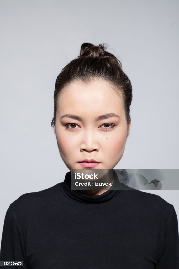 Angry asian young woman Headshot of irritated asian young woman wearing black turtleneck, staring at camera. Studio shot, grey background. Anger Stock Photo