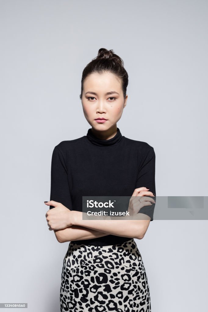 Angry asian young woman Portrait of irritated asian young woman wearing black turtleneck and leopard print skirt, standing with arms crossed and looking at camera. Studio shot, grey background. Anger Stock Photo
