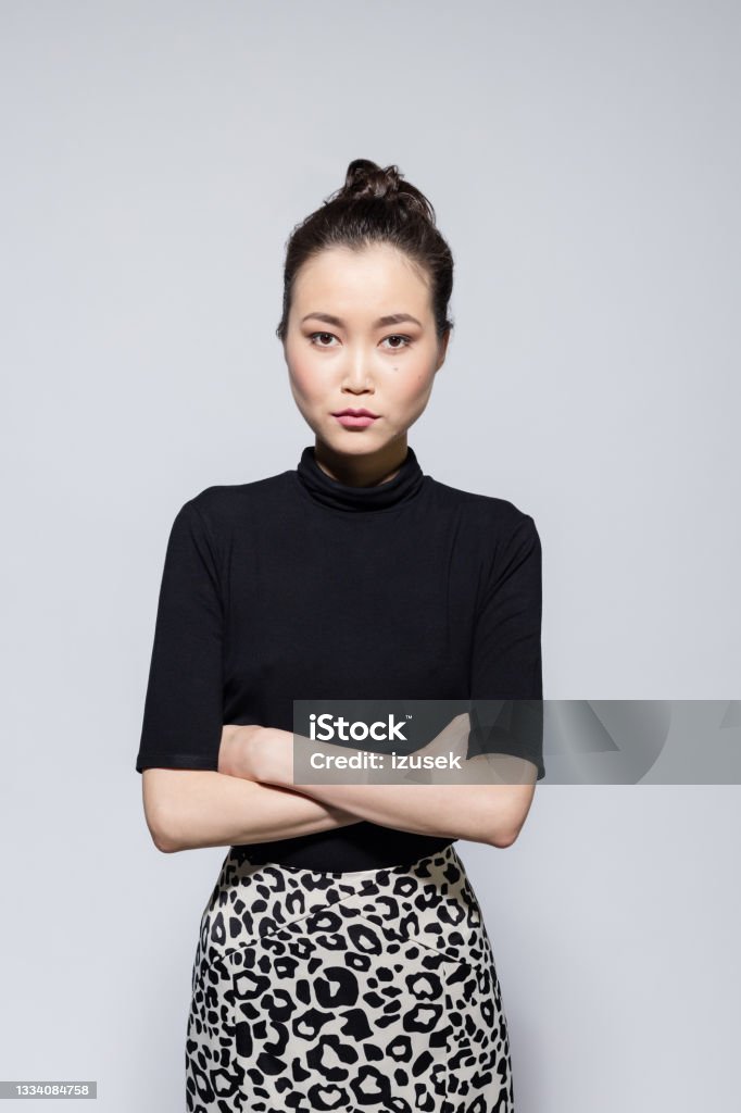 Confident asian young woman Portrait of bossy asian young woman wearing black turtleneck and leopard print skirt, standing with arms crossed and looking at camera. Studio shot, grey background. 20-24 Years Stock Photo