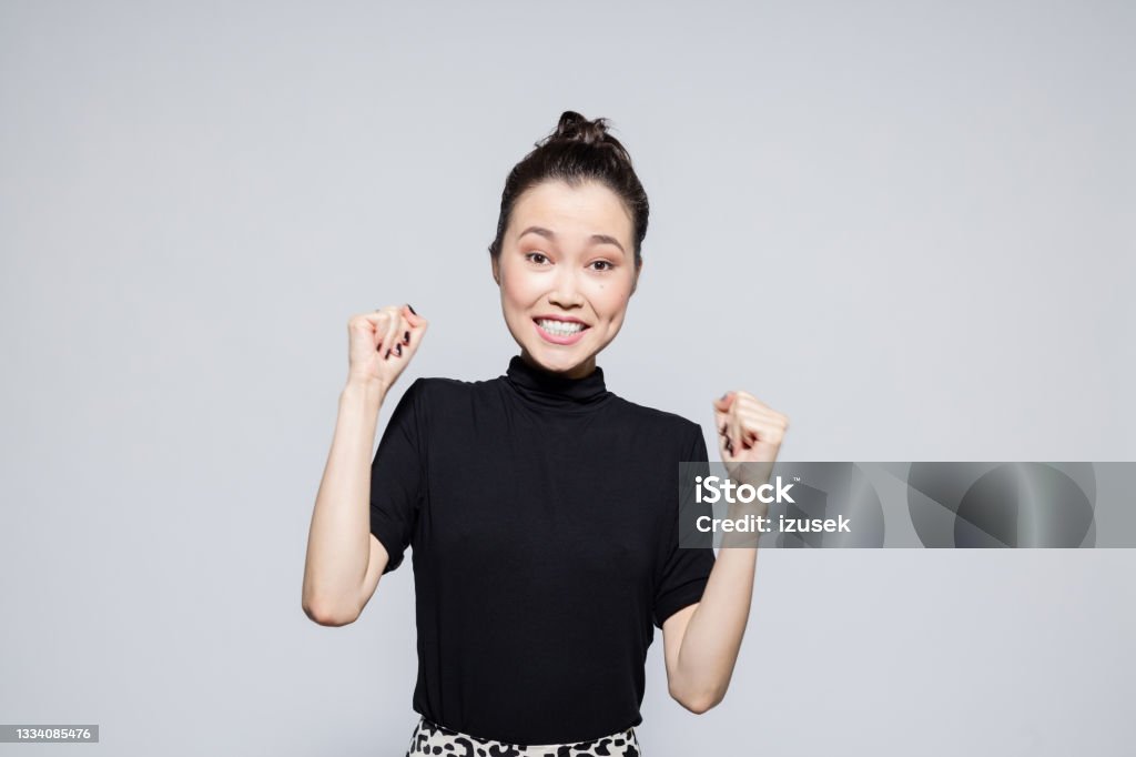 Excited asian young woman Portrait of happy asian young woman wearing black turtleneck, raising fist and laughing at camera. Studio shot, grey background. Fist Stock Photo