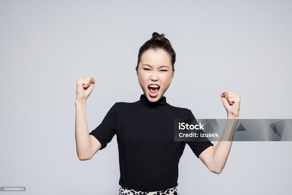 Excited asian young woman Portrait of happy asian young woman wearing black turtleneck, rising fist and shouting at camera. Studio shot, grey background. Women Stock Photo
