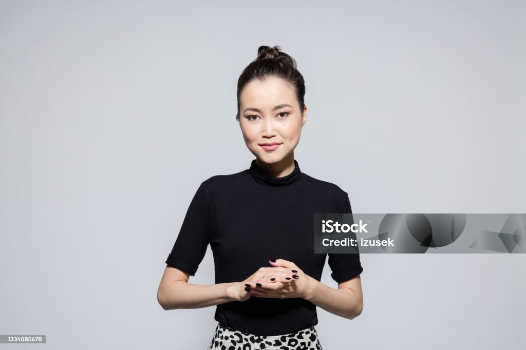 Friendly asian young woman Portrait of beautiful asian young woman wearing black turtleneck and leopard print skirt, smiling at camera. Studio shot, grey background. Mongolian Ethnicity Stock Photo