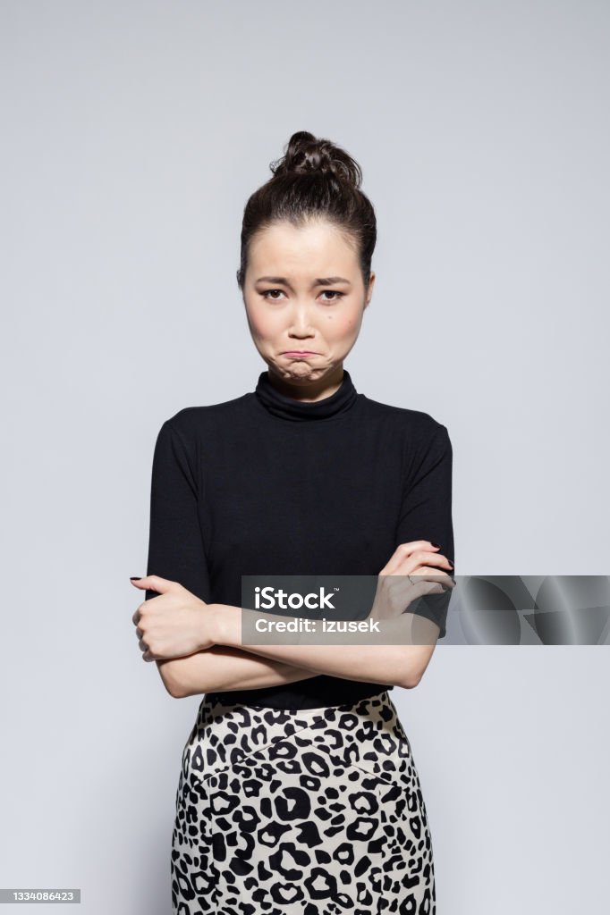 Sad asian young woman Portrait of disappointed asian young woman wearing black turtleneck and leopard print skirt, looking at camera. Studio shot, grey background. Arms Crossed Stock Photo