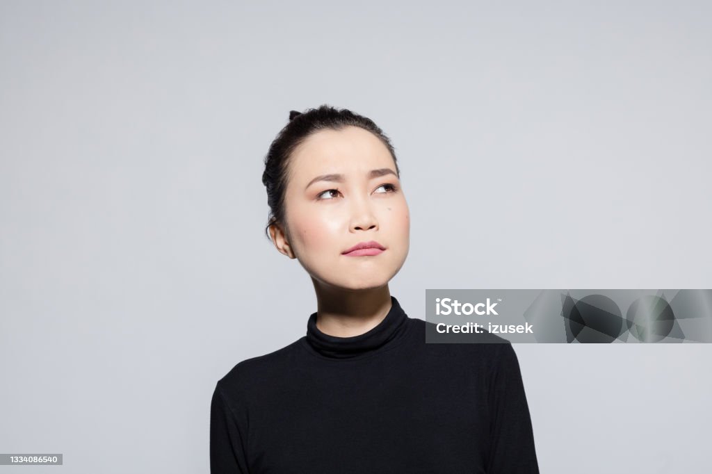 Thoughtful asian young woman Headshot of beautiful asian young woman wearing black turtleneck, looking away. Studio shot, grey background. Headshot Stock Photo