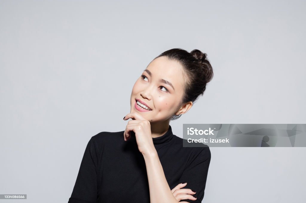 Thoughtful asian young woman Portrait of beautiful asian young woman wearing black turtleneck, looking away with hand on chin. Studio shot, grey background. Smiling Stock Photo