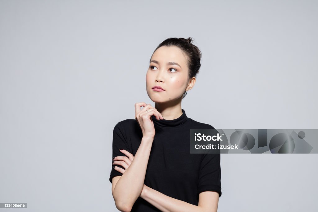 Thoughtful asian young woman Portrait of beautiful asian young woman wearing black turtleneck, looking away with hand on chin. Studio shot, grey background. Contemplation Stock Photo