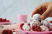 Close-up image of unrecognisable person selecting individual, freshly baked raspberry amaretti cookie from plate beside pink muslin and coffee mug, crumbs on marble effect background, focus on foreground