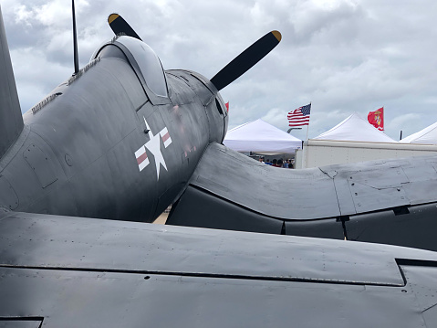 Photo of a WW2 era vintage Vought F4U Corsair plane from behind, tents, United States and Marine corps flags flying in the wind on a backdrop of cloudy sky.
