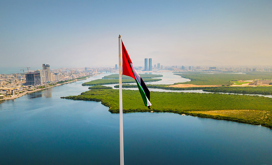 UAE national flag and Ras al Khaimah emirate aerial cityscape landmark skyline rising over the mangroves and the creek water surrounded by seaside and coastline in the United Arab Emirates aerial view.\nUAE northern emirate