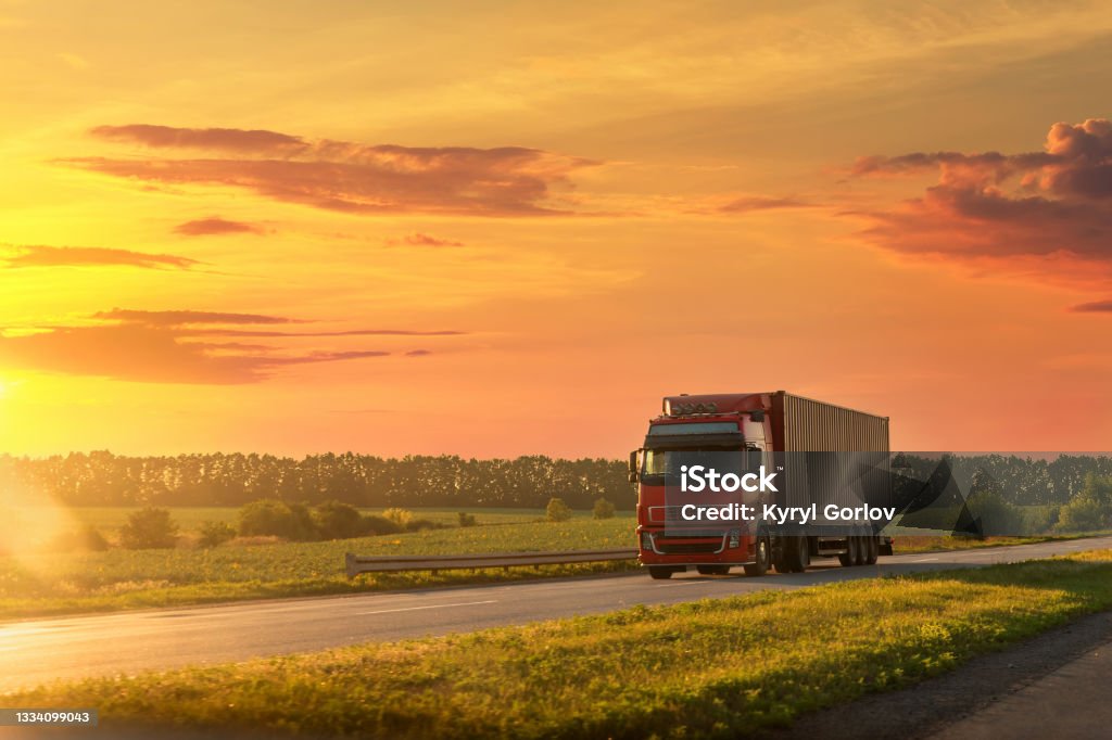 Scenic front view big long heavy semi-treailer truck with sea shipping container driving highway dramatic warm morning evening sunrise sky sunset. Cargo transport industry background concept. Truck Stock Photo