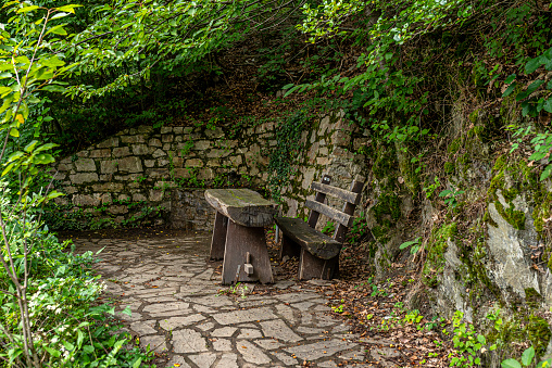 An old bench and a wooden table standing on a stone floor next to a water spring in the forest, a stone wall visible.
