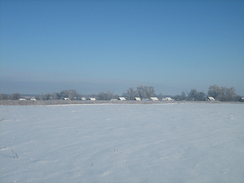 A rural flat field covered with snow on a clear frosty day. In the distance, you can see the houses of the village