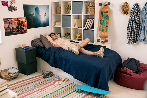 Young man lying on bed among shelf with books and skateboards while having rest and thinking