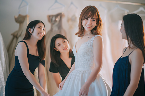 asian chinese bride trying on wedding dress surrounding by bridesmaid admiring examining the beauty of wedding gown at bridal shop