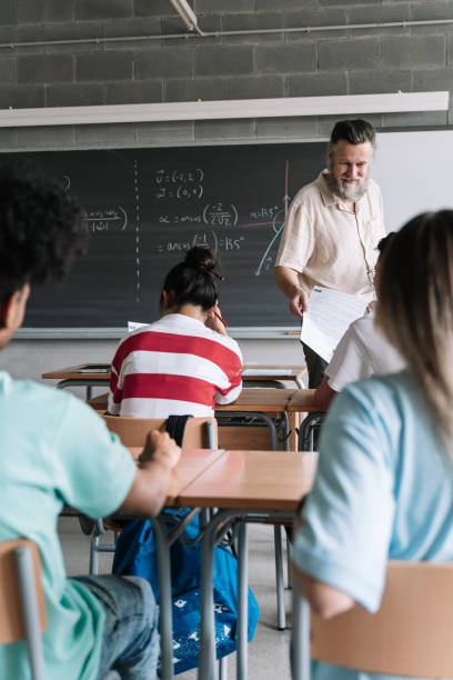 クラスの生徒に数学の試験を与えるひげを生やした教師。大学の高校の教室で多民族ティーンエイジャーの学生の背面図 - high school student test results adult student blackboard ストックフォトと画像