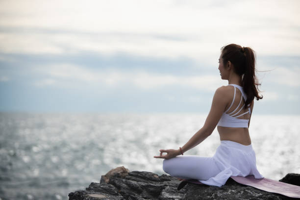 Asian young woman practicing yoga in sukhasana exercise while looking out to the sea. stock photo