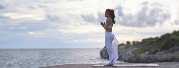 istock Asian young woman practicing yoga with sea view. Easy yoga poses for beginners. Exercise while looking out to the sea. Meditation and Strengthening Exercises with yoga. Banner size with copy space. 1334140065