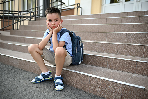 Upset worried boy with backpack sitting on the stairs by school. Bullying, loneliness, studying difficulties