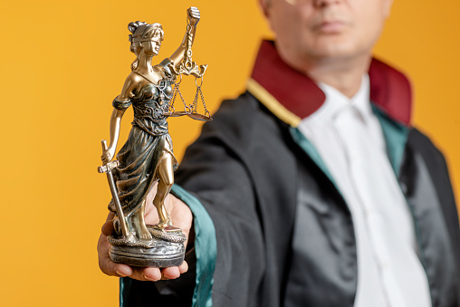Unrecognizable male lawyer is showing Lady Justice sculpture  to camera in front of yellow background