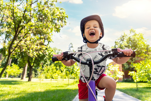 Portrait little cute adorable caucasian toddler boy in safety helmet enjoy having fun riding exercise bike in city park road yard garden forest. Child first bike. Kid outdoors sport summer activities.