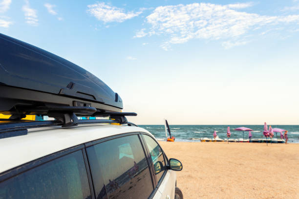 Side view of big family white suv van car with big rooftop cargo rack box at scenic sand seaside campsite camp coast of sea or ocean sky on background. Accessory rental travel trip vacation concept. vista lateral del gran coche furgoneta suv blanco de la familia con una gran caja de estante de carga en la azotea en la pintoresca arena junto al mar camping camping costa de mar o cielo oceánico en el fondo. concepto accesorio de viaje de viaje de viaje