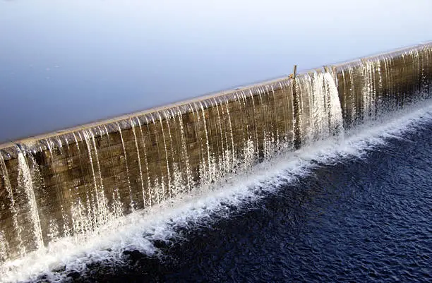 Diagonal view at a dam wall, water is overflowing seamless a long stone wall and creating a thin waterfall. Part of a lock system to create Hydro energy for a mill.