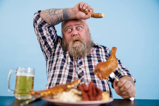 istock Tired obese man mops brows suffering from overeating at table with fat food in studio 1334171713