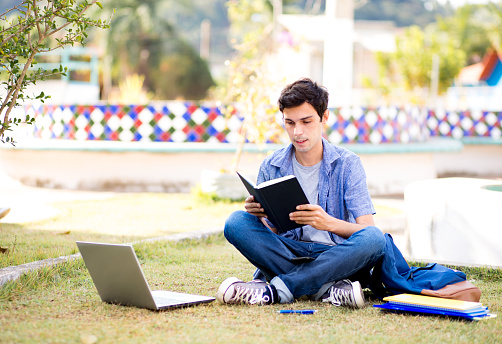 Young college student sitting on college campus reading a book and using laptop.