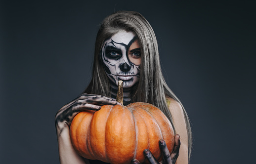 Young woman with long hair and skull face art holding big pumpkin and looking at camera on Halloween party against dark gray background