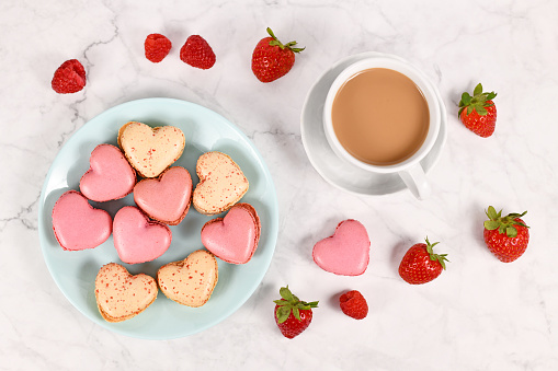 Pink and beige heart shaped French macaron sweets next to coffee cup and strawberry and raspberry fruits
