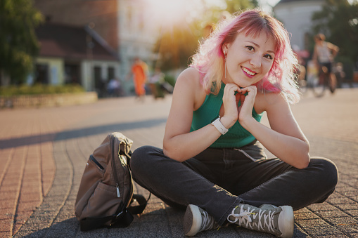Stylish young smiling woman with multicolored pink hair sitting on the pavement in a sunny city holding hands in the heart shape