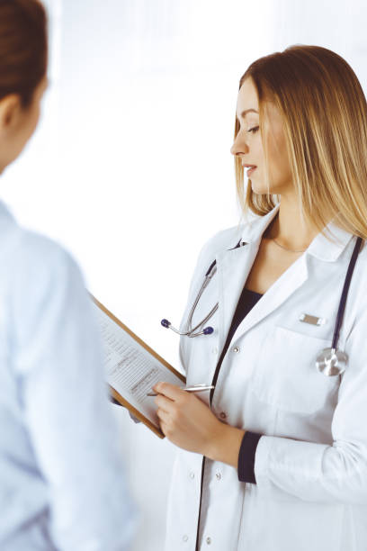 Young woman-doctor and her patient are discussing patient's current health examination, while standing together in a hospital office. Female physician is writing some marks, using a clipboard. Perfect medical service in a clinic. Medicine concept. la joven doctora y su paciente están discutiendo el examen de salud actual del paciente, mientras están juntos en una oficina del hospital. la médica está escribiendo algunas marcas, usando un portapapeles. servicio médico perfecto en una clínica. co