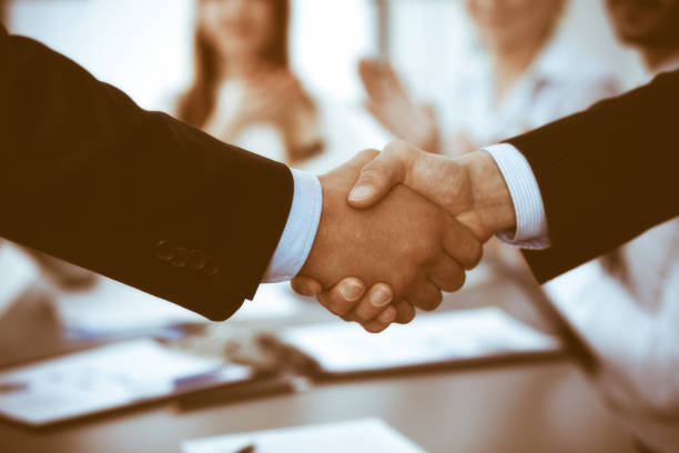 istock Business people shaking hands at meeting while theirs colleagues clapping and applauding. Group of unknown businessmen and women in modern white office. Success teamwork, partnership and handshake concept 1334248318